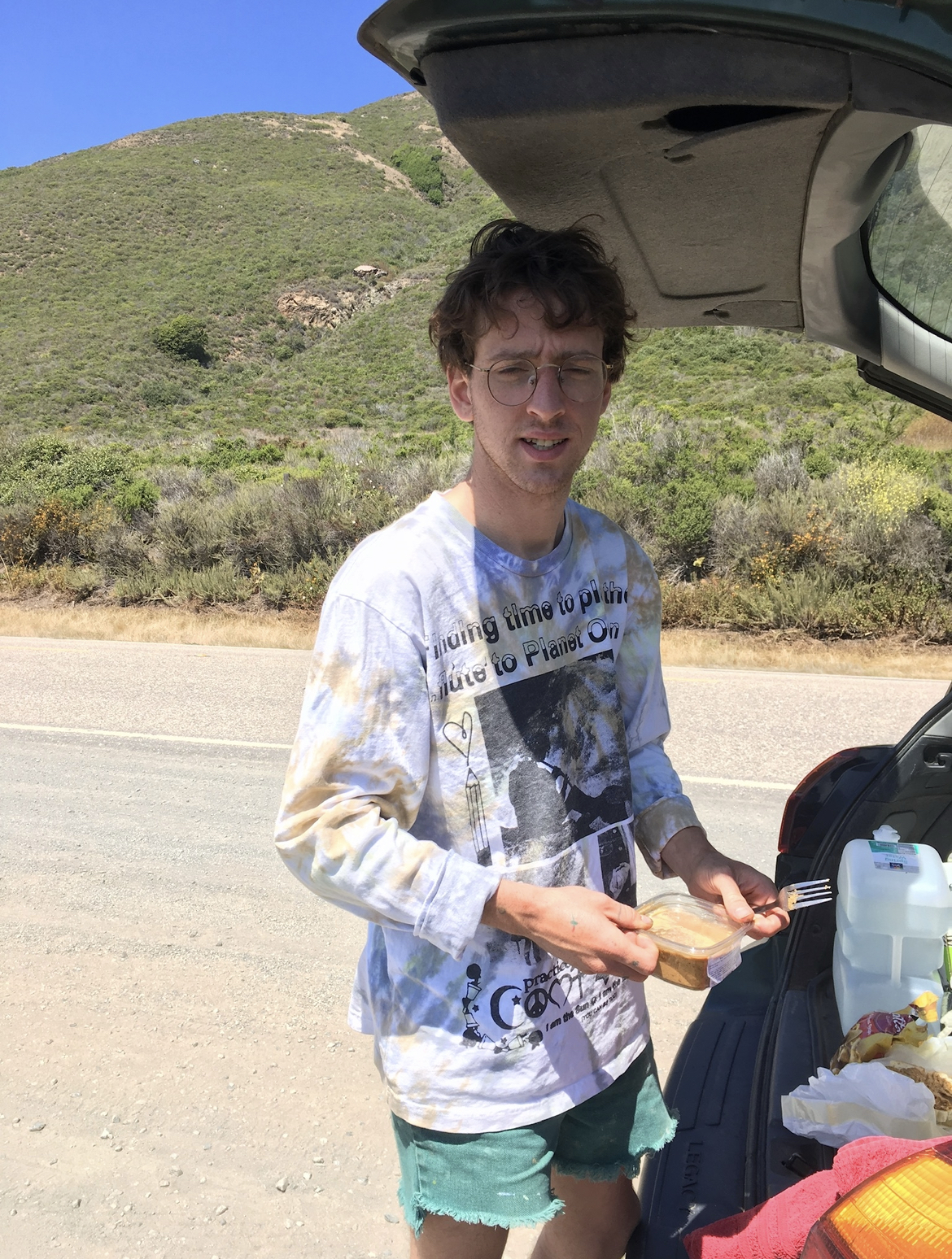 A photo of me, a white man with messy brown hair and round glasses, wearing a long sleeve tie dye t-shirt and green shorts made from cut off jeans. I am standing on the side of Highway 1 in Big Sur, California. Behind me is a verdant green hill and a small portion of clear blue sky. I'm standing behind the open hatch of a hatchback vehicle, holding a container of almond butter and a fork.